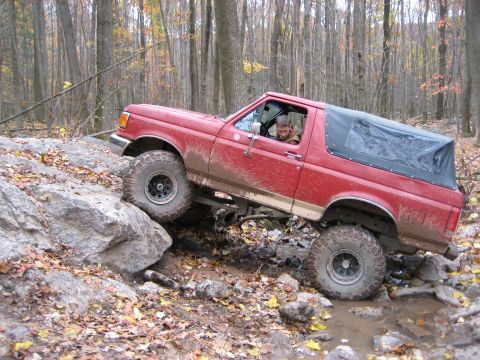 1990 Ford Bronco - A red 1987 Ford Bronco tackles a challenging off-road course with large rocks and mud.