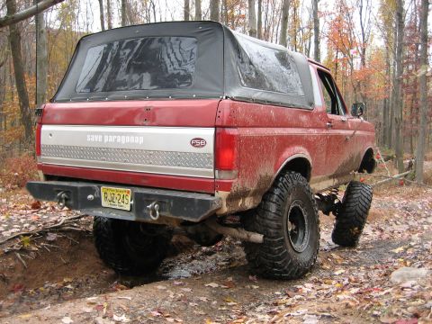 1990 Ford Bronco - This red 1990 Ford Bronco, equipped for off-roading, navigates a muddy trail surrounded by autumn trees.