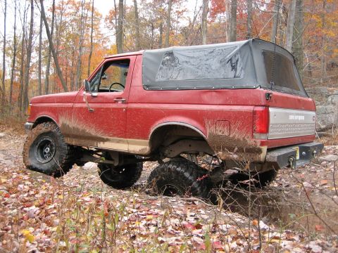 1990 Ford Bronco - A red 1990 Ford Bronco navigates a muddy trail in a wooded area during autumn.