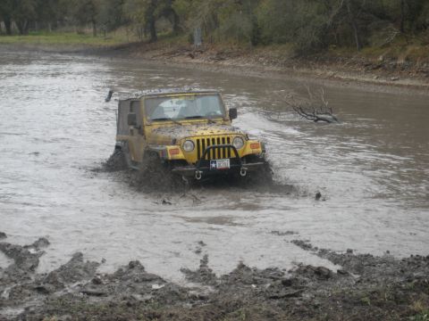2001 Jeep Wrangler - A yellow Jeep Wrangler navigates a muddy, water-filled terrain, kicking up spray as it moves forward.