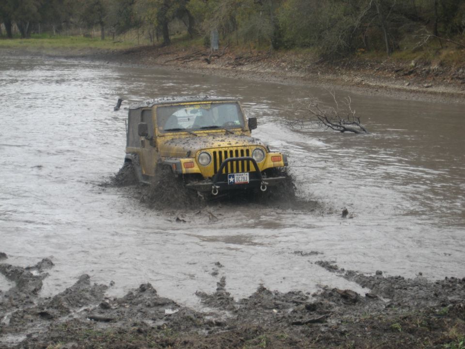 Yellow Jeep Wrangler splashing through muddy water, front view. License plate visible