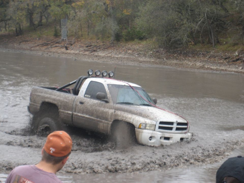 White Dodge Ram pickup truck with roll bar and spotlights stuck in mud, viewed from the front-right