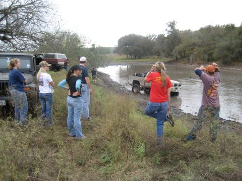 2001 Jeep Wrangler - Several people stand on a muddy bank observing a truck navigate through a shallow, murky waterway.
