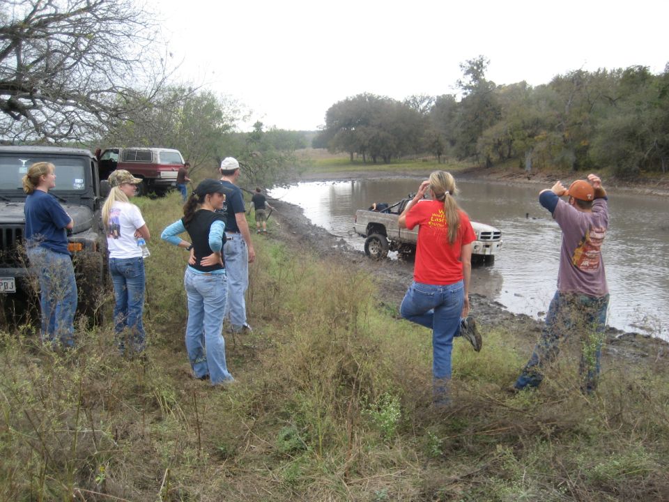 Group of people watching a white pickup truck drive through a muddy body of water