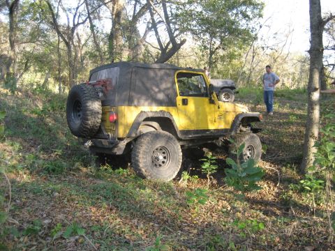 2001 Jeep Wrangler - A yellow 2001 Jeep Wrangler is parked on a wooded trail, with another vehicle and a person in the background.