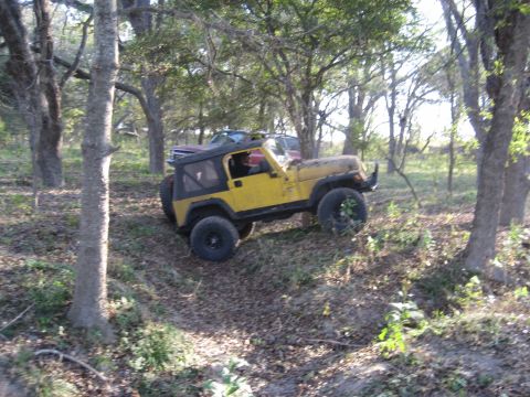 2001 Jeep Wrangler - A yellow Jeep Wrangler navigates a wooded trail, with another vehicle partially visible behind it.