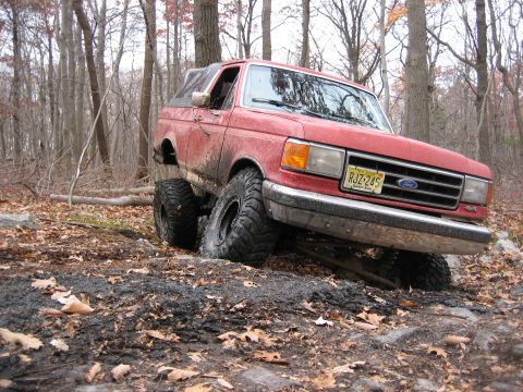 1990 Ford Bronco - A red Ford Bronco navigates a challenging off-road course with its tires deep in mud and rocks.