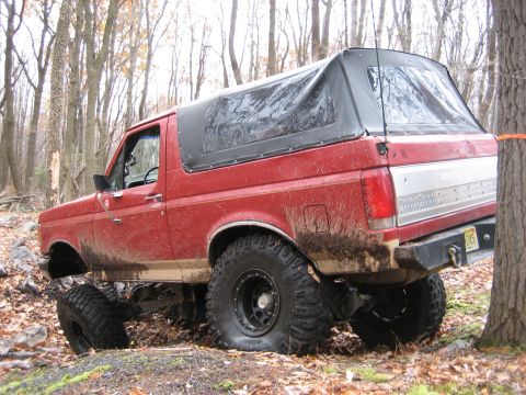 1990 Ford Bronco - Red 1990 Ford Bronco navigates a wooded trail with mud splattered on its body.