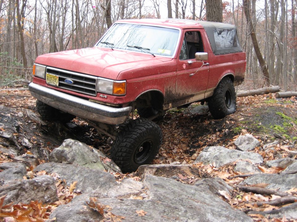 Red 1990 Ford Bronco climbing over rocks on a wooded trail