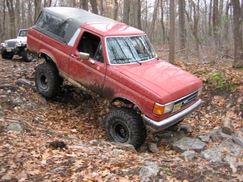 1990 Ford Bronco - This red SUV is navigating a challenging rocky trail in a wooded area, with another vehicle following behind.