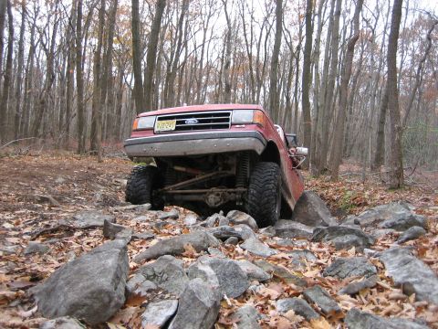 1990 Ford Bronco - This red SUV navigates a rocky trail through a dense forest during autumn.
