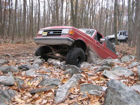 1990 Ford Bronco - A red 1990 Ford Bronco is shown off-roading through a rocky forest trail with another vehicle in the background.