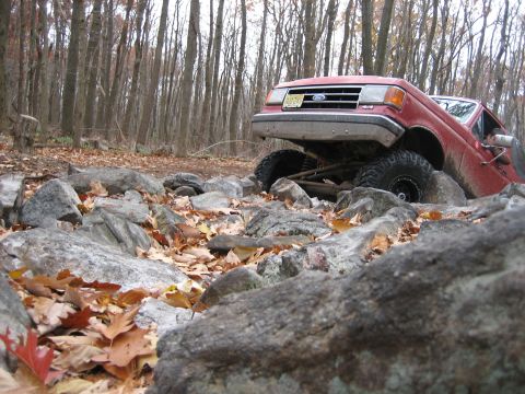 1990 Ford Bronco - A red 1990 Ford Bronco tackles a challenging rocky trail in a wooded area.
