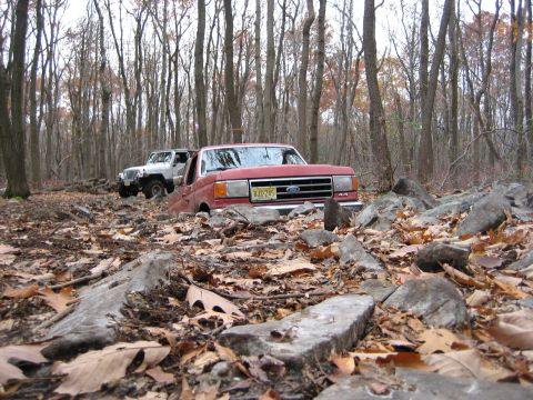 1990 Ford Bronco - Two off-road vehicles, a red Ford Bronco and a white Jeep, are seen in a wooded area with rocky terrain.