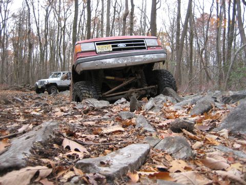 1990 Ford Bronco - A red Ford Bronco tackles a challenging off-road course with a white Jeep in the background.