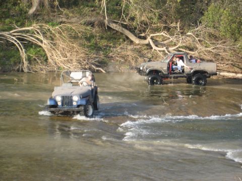2001 Jeep Wrangler - Two off-road vehicles navigate a shallow river, with a blue Jeep CJ-7 in the foreground and a lifted pickup truck following.