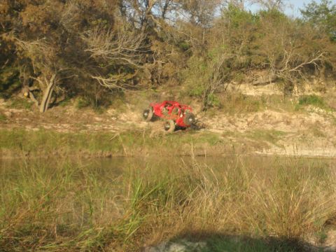 2001 Jeep Wrangler - A red off-road vehicle is positioned on a dirt bank next to a body of water, surrounded by trees and vegetation.