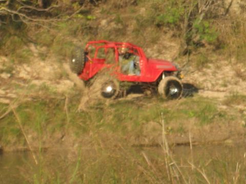 2001 Jeep Wrangler - A red Jeep Wrangler navigates a dirt path near a body of water, kicking up some dust.