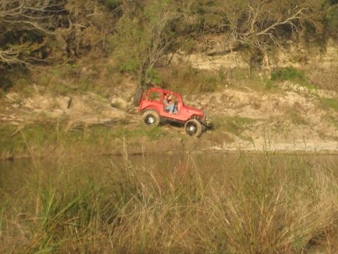 2001 Jeep Wrangler - A red Jeep Wrangler navigates a steep, grassy embankment near a calm body of water.