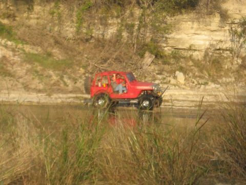 2001 Jeep Wrangler - A red Jeep Wrangler navigates through shallow water, with a rocky embankment in the background.
