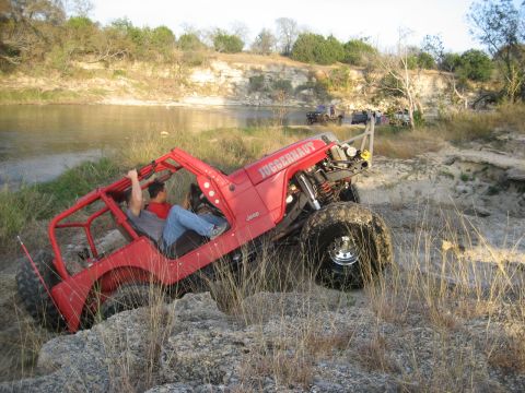 2001 Jeep Wrangler - A heavily modified Jeep navigates a rocky terrain near a body of water, with other vehicles visible in the distance.