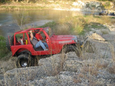 2001 Jeep Wrangler - A red Jeep Wrangler navigates rough terrain by a river, with other vehicles visible in the background.