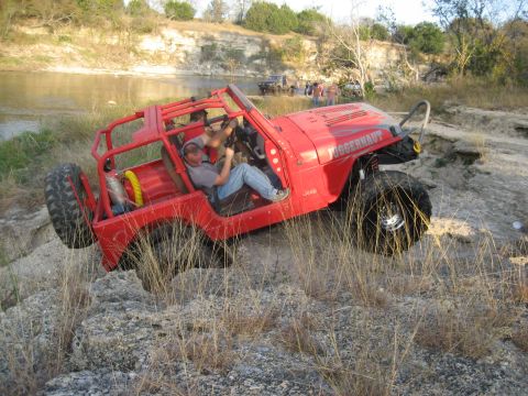 2001 Jeep Wrangler - A red Jeep navigates a rough, off-road course by the water, with other vehicles and people in the background.