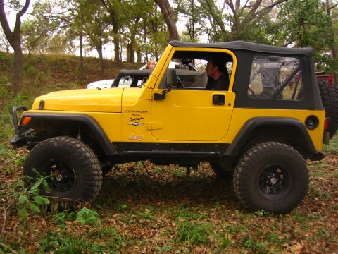 2001 Jeep Wrangler - Side view of a yellow 2001 Jeep Wrangler 4.0L Sport with a black soft top, parked in a wooded area.