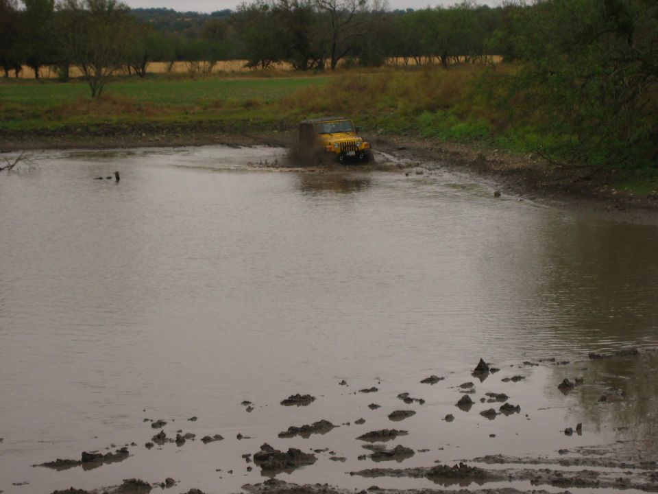 Yellow 2001 Jeep Wrangler driving through a muddy puddle with trees and fields in the background