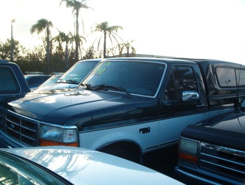1995 Ford F-150 - A dark green and white pickup truck with a camper shell is parked in a lot with other vehicles and palm trees.