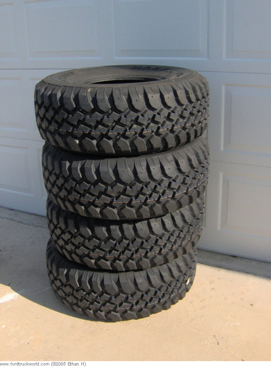 Stack of four new mud-terrain off-road tires on a driveway in front of a garage door