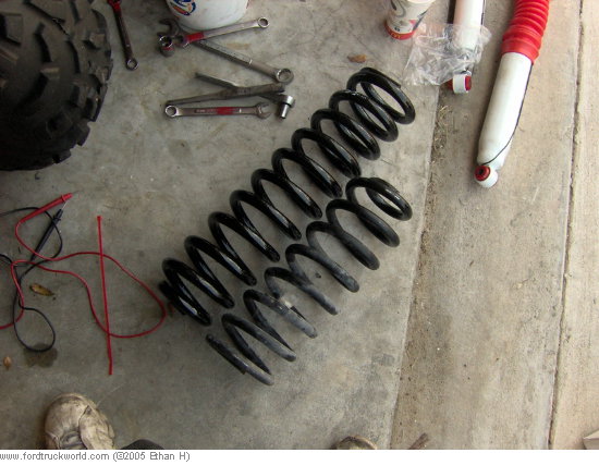 Two black coil springs and a red-topped shock absorber lie on a concrete floor next to a tire and tools