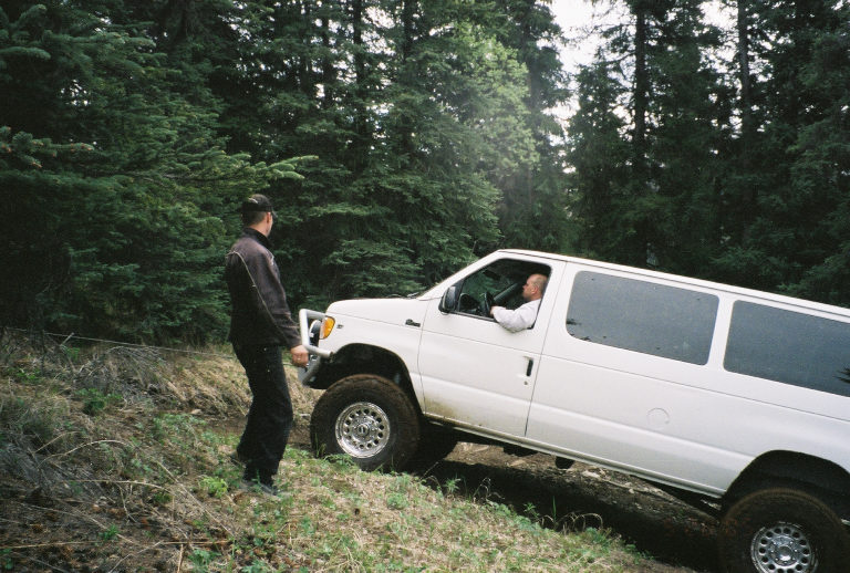 White Ford E350 van with off-road tires on a dirt path, driver visible inside