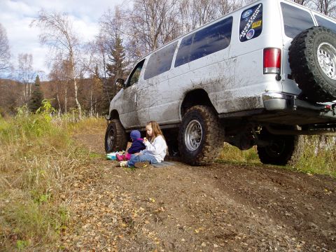 2000 Ford E350 - Low 3/4-rear view of a lifted white Ford full-size van on 4x4 running gear, lower body and windows coated in dried mud, parked on a muddy t…