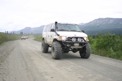 2000 Ford E350 - A modified 2000 Ford E350 van with a snorkel and oversized tires navigates a dusty gravel road in a mountainous, wooded area.