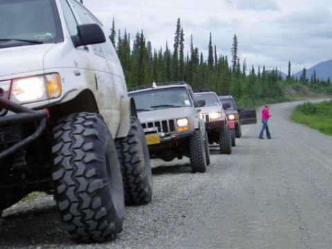 2000 Ford E350 - A line of off-road vehicles drives down a gravel road with trees and mountains in the background.