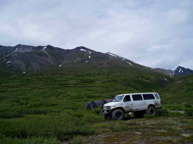 White Ford E350 van with large off-road tires parked in a grassy field with mountains in the background