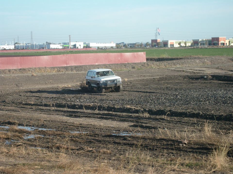 1989 Ford Bronco driving through a muddy field with buildings in the background