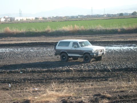 1989 Ford Bronco - Wide shot of a lifted white-over-grey Ford Bronco with hardtop driving across a muddy, rutted field with standing water, occupants visible…