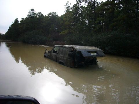 1989 Jeep Cherokee - A dark 1989 Jeep Cherokee is partially submerged in muddy water, its rear hatch open, as it appears to be stuck.