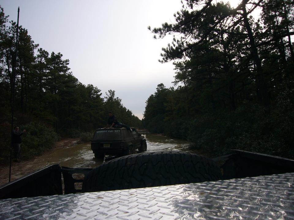 off-road vehicle driving through mud, another vehicle behind it, trees on both sides