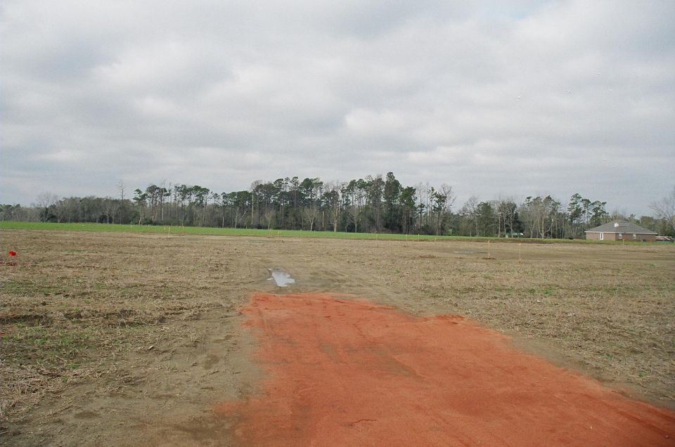 Construction site with bare ground, orange dirt, and trees in the background under a cloudy sky. A house is visible on the right