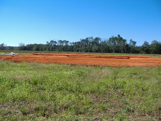 Construction site with cleared land and dirt mounds, trees in background under a clear blue sky