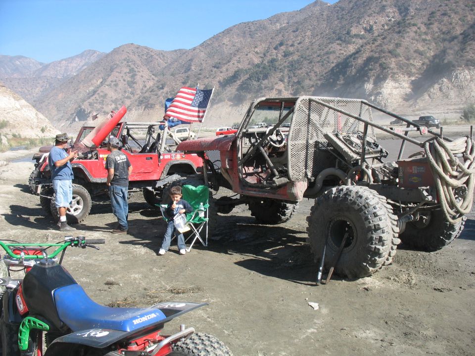Red tube-frame rock buggy on huge tires and a red Jeep at a desert off-road staging area