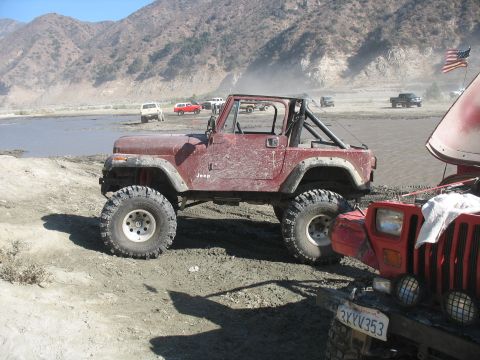 1987 Ford Bronco - A red Jeep Wrangler with large off-road tires sits on a dusty terrain, with another vehicle partially visible in the foreground.