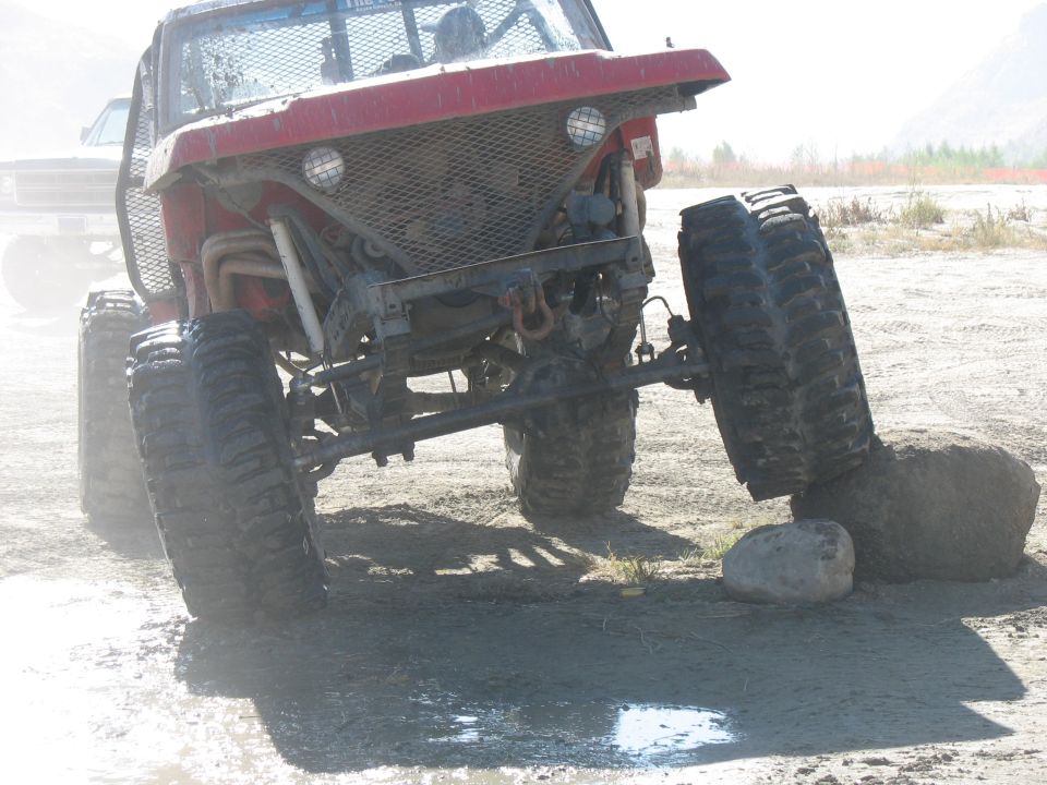 red off-road vehicle with large tires, one tire on a rock, another vehicle behind it