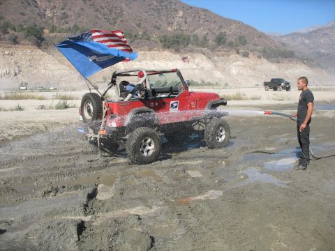 1987 Ford Bronco - A red Jeep Wrangler is being hosed down in a muddy, outdoor environment with mountains in the background.