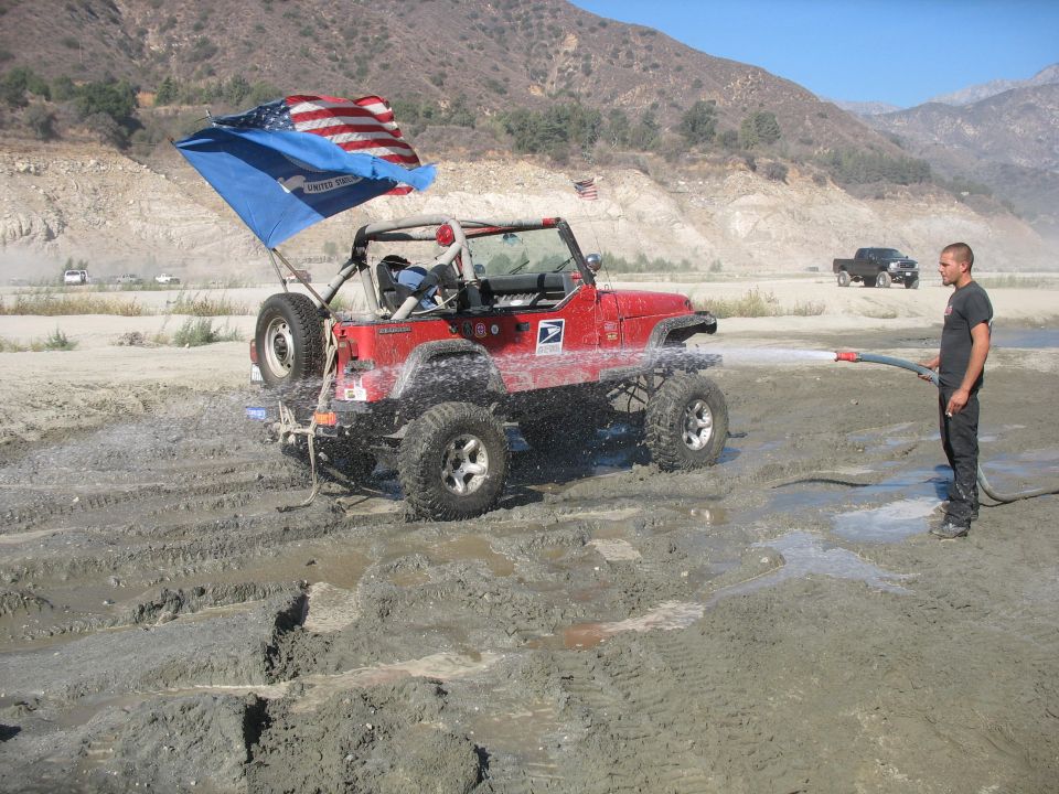 Red Jeep Wrangler with American flag, being sprayed with water in a muddy area