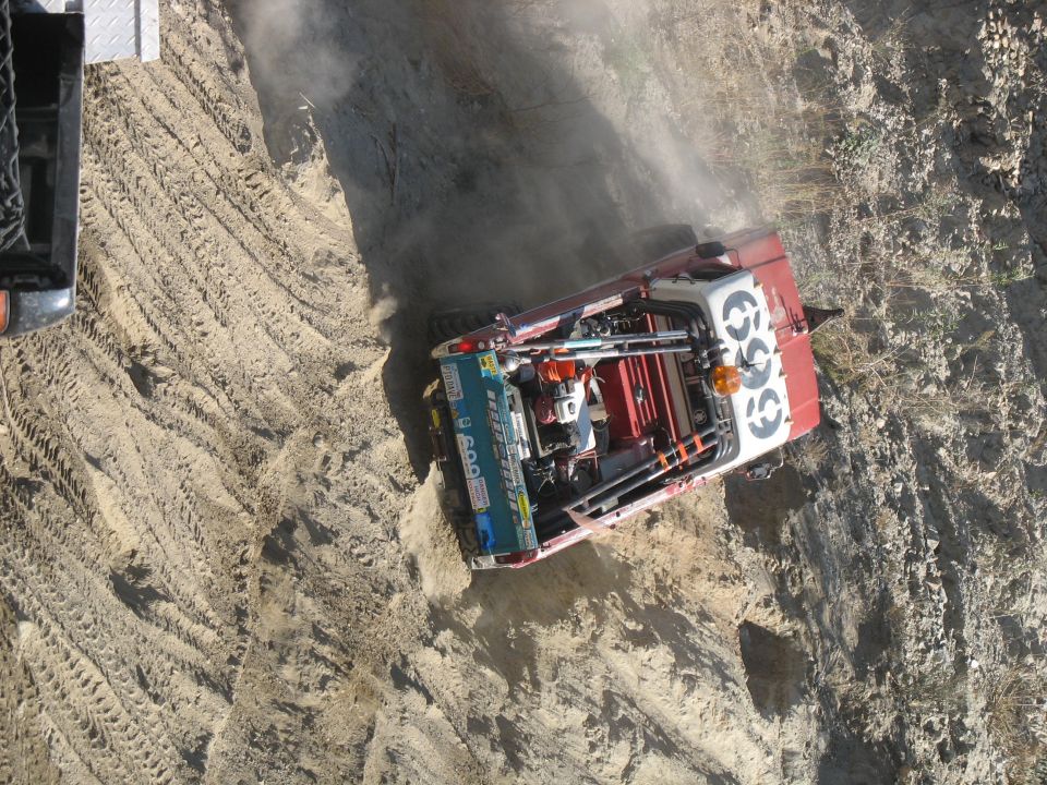 Off-road truck with roll cage and large tires on a dirt track. Another vehicle is partially visible on the left