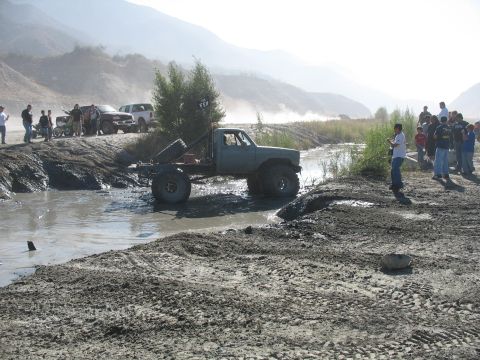 1987 Ford Bronco - A heavily modified pickup truck navigates a muddy off-road course with spectators watching.